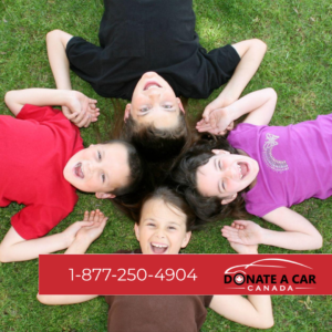 Four laughing children wearing solid coloured shirts are lying face up, head to head, in a field of green grass. Hope Donate a Car Canada