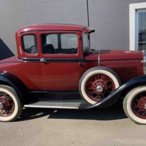 Passenger side view of donated burgundy 1931 FORD MODEL A with red hubs and white wall tires parked in front of a gray wall awaiting auction sale so that donation can be sent to the donor's charity of choice.